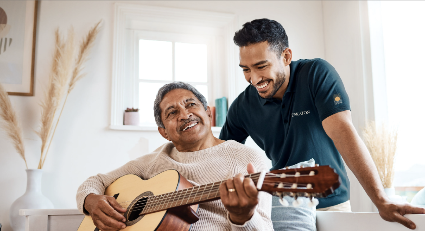 An older man playing an acoustic guitar smiles while a younger man stands beside him, also smiling, in a bright, cozy room with natural light.